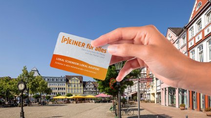 Hand hält den Stadtgutschein „Peiner für alles“ vor dem Peiner Marktplatz mit historischen Gebäuden, Cafés und Sonnenschirmen an einem sonnigen Tag.