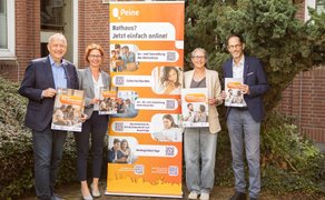 Vier Personen stehen vor einem Gebäude neben einem orangefarbenen Aufsteller zum digitalen Rathaus der Stadt Peine und halten Informationsmaterial in den Händen.