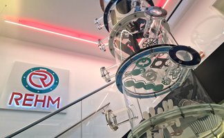 Transparente Kunststoffsäule mit technischen Dichtungen und Metallteilen vor einer Wand mit beleuchtetem REHM-Logo