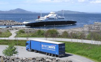 Lkw mit Aufschrift „FRITZ BENTE Spezial- & Großraumtransporte“ auf einer Straße am Wasser mit einer Fähre und Inseln im Hintergrund.