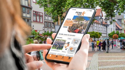 Person nutzt die Stadt-App „Peine2Go“ auf dem Smartphone in der belebten Peiner Innenstadt mit Fachwerkhäusern und Fußgängerzone im Hintergrund.