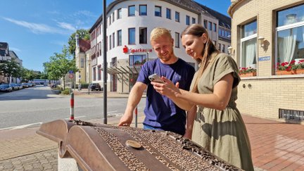 Zwei Teilnehmende nutzen ein Smartphone und eine Infostation in der Innenstadt von Peine während einer digitalen Stadtrallye.“
