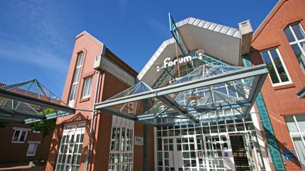 Eingangsbereich eines modernen Backsteingebäudes mit Glasvordach und dem Schriftzug „Forum“.
