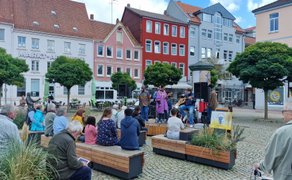 Open-Stage-Konzert auf dem Marktplatz in Peine mit Live-Band auf einer kleinen Bühne, sitzenden Zuschauerinnen und Zuschauern auf Holzbänken und farbenfrohen Fassaden der Innenstadt im Hintergrund.