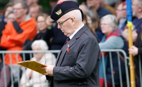 Älterer Mann in Uniform mit Mütze hält ein gelbes Klemmbrett vor einer Menschenmenge hinter Absperrungen