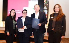 Vier Personen stehen bei der Übergabe von Förderbescheiden des Programms „Zukunftsräume Niedersachsen“ vor einem Bildschirm mit Präsentationsfolie in einem historischen Saal.