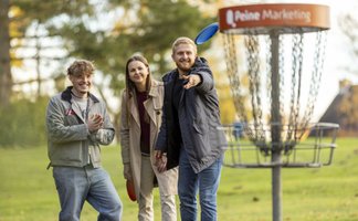 Drei junge Erwachsene spielen Discgolf im Grünen in Peine und werfen eine Frisbee-Scheibe auf einen Discgolf-Korb.