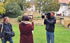 Eine Frau richtet ein Tablet auf eine Parkanlage mit alten Gemäuern und wird dabei von Kameramann und Tontechniker aufgenommen.