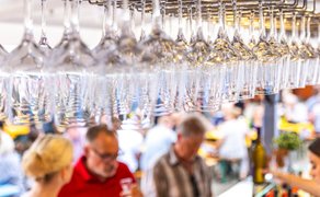 Reihenweise aufgehängte Weingläser über einem belebten Ausschankstand beim Peiner Weinfest, mit Gästen im Hintergrund.