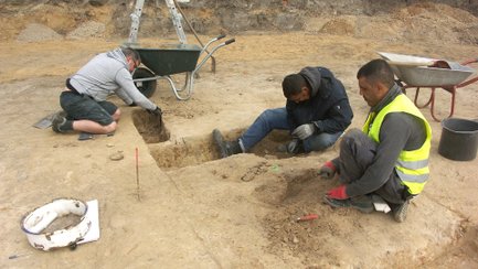 Drei Personen graben in einer archäologischen Ausgrabungsstätte in einer rechteckigen Grube im Boden.