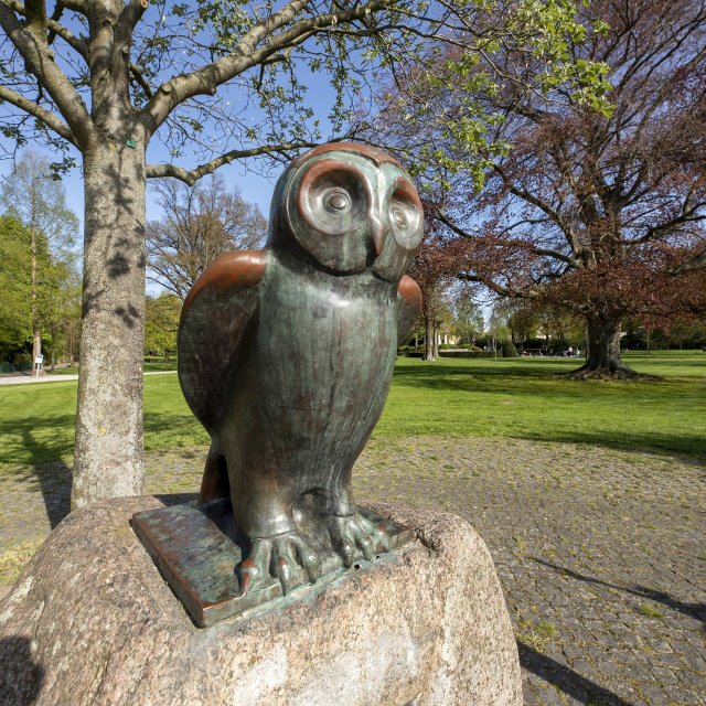Bronzeskulptur einer Eule im Stadtpark Peine mit grüner Parklandschaft und alten Bäumen im Hintergrund.