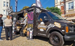 Ein schwarzer Foodtruck steht auf einem Platz, mehrere Menschen warten vor der geöffneten Verkaufsklappe.