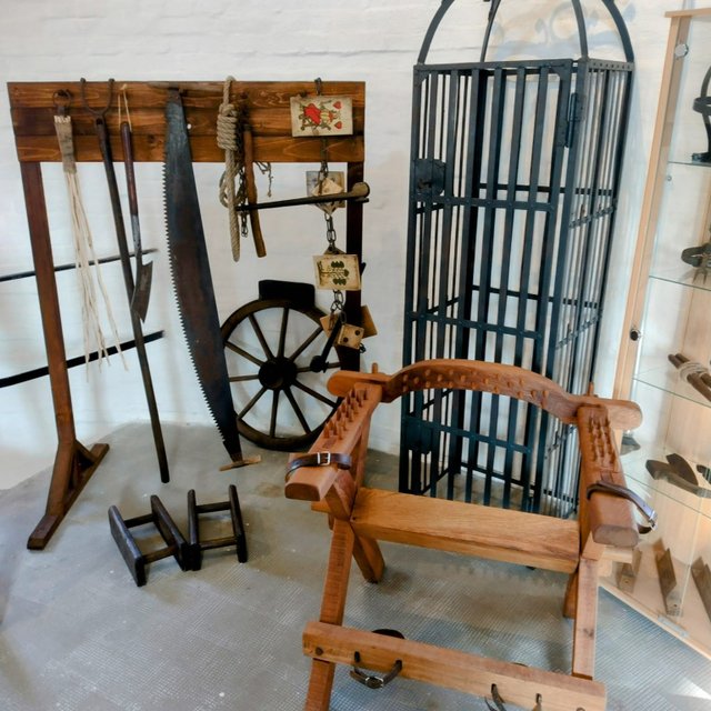 Ausstellung historischer Folterinstrumente mit Holzgestell, Säge, Seilen, Streckbank und Eisenkäfig in einem Museumsraum, die die Methoden der Justiz zur Zeit der Hexenverfolgung veranschaulichen.