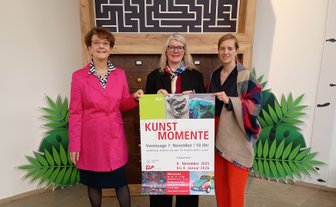 Drei Frauen stehen vor einer Holzwand mit Labyrinth-Optik und halten ein Plakat mit der Aufschrift „KUNST MOMENTE“ und „KiP“ in den Händen.