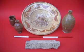 Vier archäologische Keramikgefäße und ein verziertes Steinfragment auf rotem Untergrund.