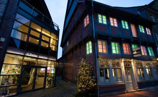 Beleuchtetes Fachwerkhaus und modernes Gebäude mit Glasfassade bei Dämmerung, geschmückt mit Weihnachtsbeleuchtung und Baum mit Lichterkette.