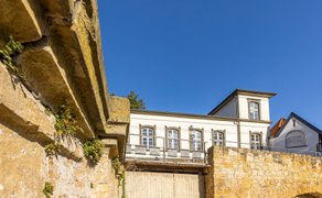 Eine historische Natursteinmauer und ein gemauerter Torbogen befinden sich im Vordergrund. Dahinter steht ein weißes Gebäude mit hohen Fenstern unter einem klaren blauen Himmel.