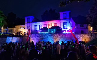 Menschen mit leuchtenden Funkkopfhörern feiern nachts vor einer bunt illuminierten historischen Kulisse im Peiner Burgpark.