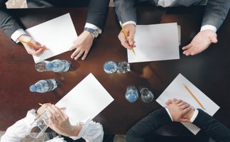 Draufsicht auf ein geschäftliches Meeting: Mehrere Personen sitzen an einem Tisch, halten Stifte über leere Blätter, daneben stehen Wasserflaschen und Gläser.