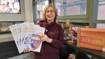 Person mit dunkelrotem Pullover hält mehrere Exemplare des Stadtmagazins "Stadt Land Leute" in der Hand, im Hintergrund eine orangefarbene Genussbox mit der Aufschrift "Bierliebe".