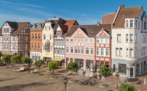 Historischer Marktplatz in Peine mit gepflastertem Platz, Bäumen und einer Reihe denkmalgeschützter Fachwerk- und Altstadthäuser unter blauem Himmel.