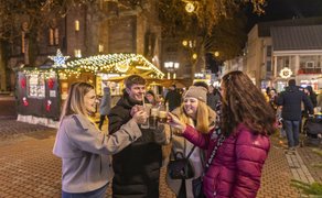 Vier Personen stoßen mit Getränken auf einem festlich beleuchteten Weihnachtsmarkt an, im Hintergrund sind Stände und weitere Besucher zu sehen.