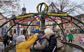 Mehrere Menschen stehen um einen Brunnen auf einem Marktplatz, der mit grünen Girlanden und bunten Ostereiern geschmückt ist. Im Vordergrund helfen zwei Erwachsene einem Kind dabei, eine Dekoration an der Girlande zu befestigen. Im Hintergrund bewegen sich weitere Besucher über den Platz, darunter Familien mit Kindern. Rundherum sind Bäume sowie historische Gebäude zu sehen.