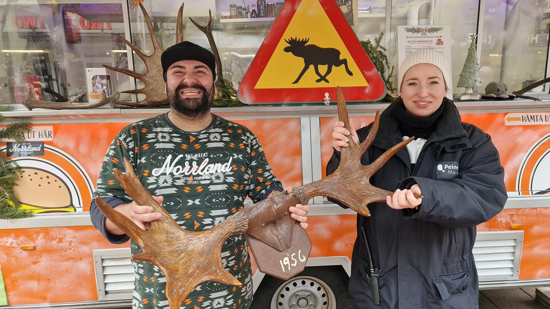 Ein Mann und eine Frau halten gemeinsam ein großes Geweih mit der Jahreszahl „1956“, im Hintergrund ein Warnschild mit Elch-Symbol an einem Foodtruck.