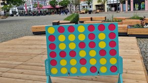 Großes, freistehendes Vier-gewinnt-Spiel mit roten und gelben Spielsteinen auf einer Holzplattform in der Peiner Innenstadt, im Hintergrund Sitzmöbel, Bepflanzung und Fachwerkhäuser.