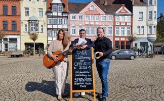 Drei Personen stehen auf dem Historischen Marktplatz in Peine vor bunten Fachwerkhäusern; eine Frau hält eine Gitarre, ein Mann ein Tamburin, in der Mitte steht eine Tafel mit der Aufschrift „Herzlich Willkommen zur Open Stage 2026“, die auf das Musik- und Kulturangebot im Freien hinweist.