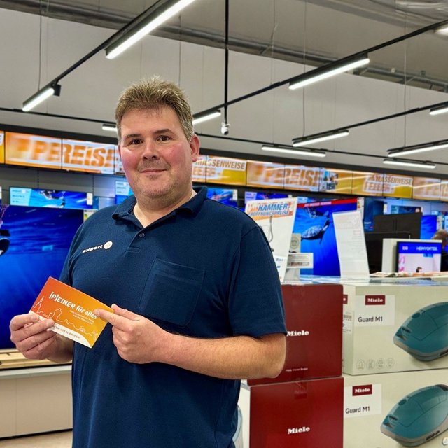 Ein Mitarbeiter steht in einem Elektrofachmarkt und hält einen „[P]EINER für alles“-Gutschein in der Hand. Im Hintergrund sind Fernseher und weitere Elektrogeräte ausgestellt.