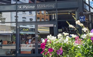 Eingang der Tourist-Information Peine im Gebäude „Peine Marketing“ mit Glasfassade und Beschilderung. Im Vordergrund stehen blühende Blumen, im Hintergrund sind Souvenirs und Informationsmaterial im Innenraum sichtbar.