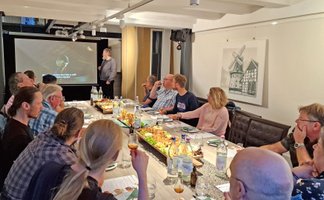 Eine Gruppe von Menschen sitzt gemeinsam an einem Tisch mit Käseplatte und Gläsern und verfolgt eine Präsentation.