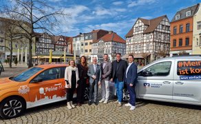 Sechs Personen stehen zwischen einem orangefarbenen Auto mit der Aufschrift „Peine“ und einem silbernen Fahrzeug mit dem Logo „Volksbank BRAWO“ vor Fachwerkhäusern auf einem gepflasterten Platz.