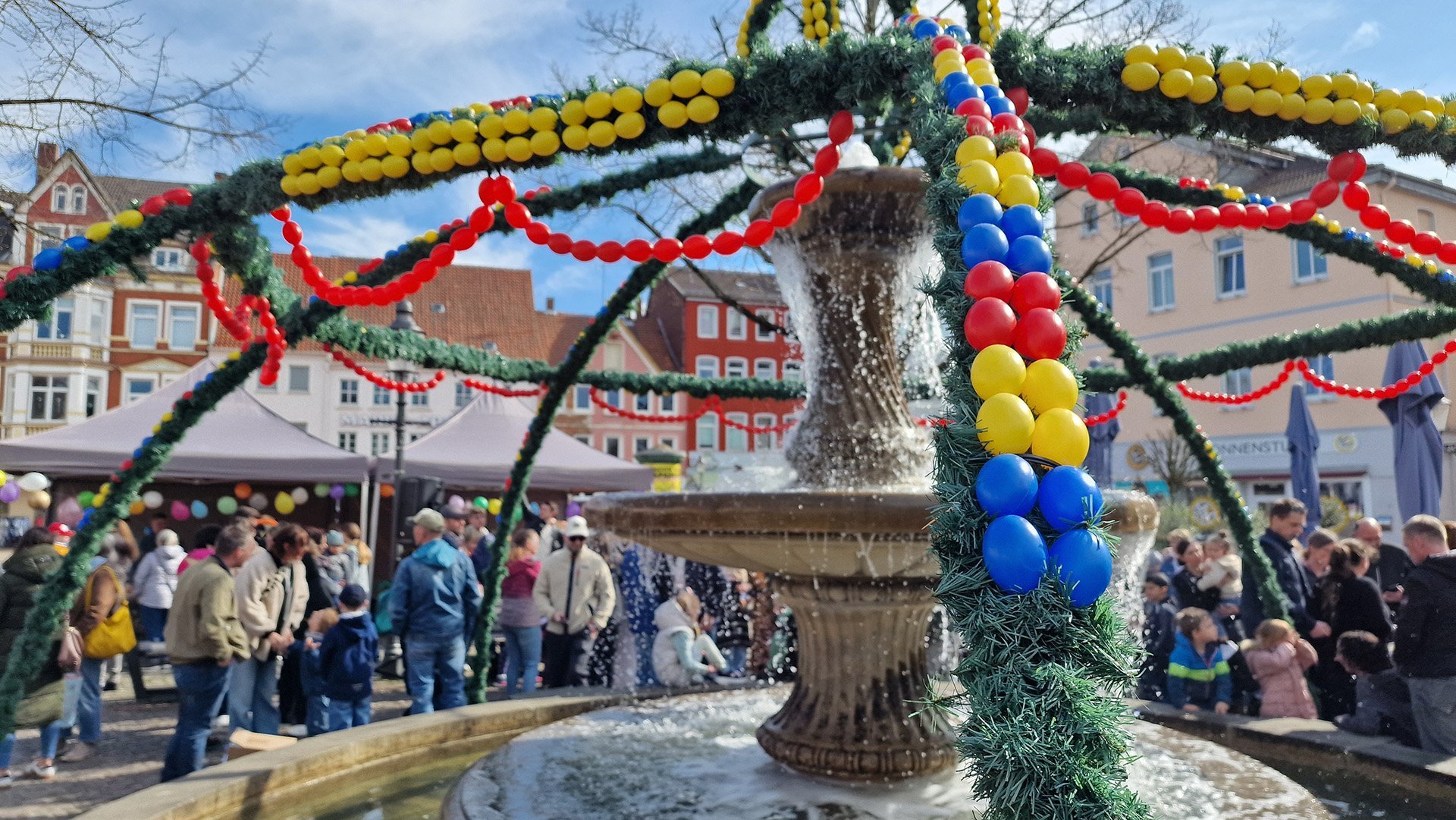 Der Osterbrunnen auf dem Peiner Marktplatz ist mit grünen Girlanden und bunten Ostereiern geschmückt, während sich viele Menschen rund um den Brunnen versammeln.