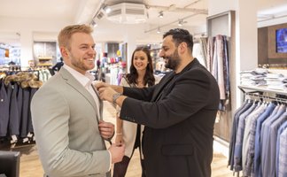 Zwei Männer stehen in einem Bekleidungsgeschäft, während einer dem anderen ein Sakko anpasst; im Hintergrund lächelt eine Frau und weitere Kleidung hängt ordentlich auf Ständern.