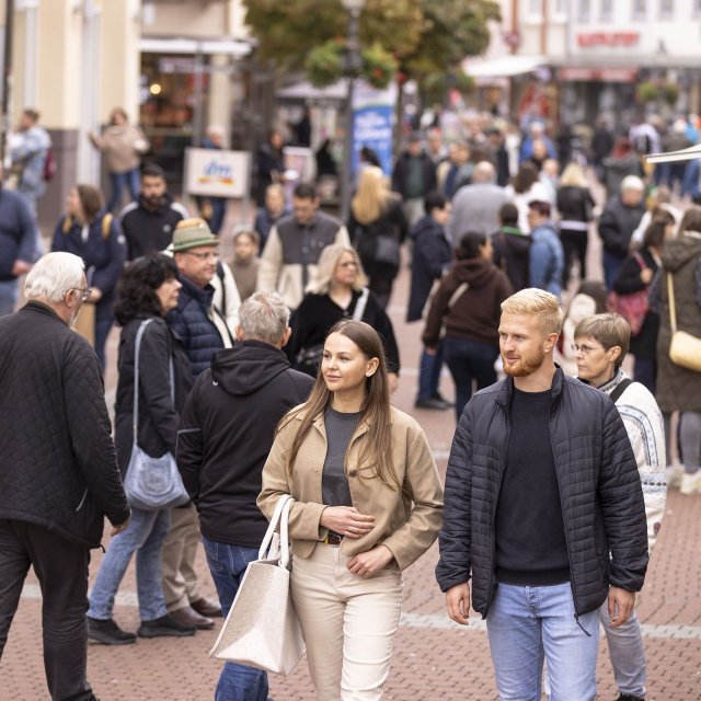 Belebte Fußgängerzone in der Peiner Innenstadt mit zahlreichen Passanten unterschiedlichen Alters, die zwischen Geschäften flanieren und einkaufen. Im Vordergrund gehen zwei junge Erwachsene durch das geschäftige Treiben der Einkaufsstraße.