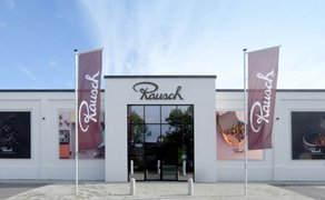 Fassade eines weißen Gebäudes mit großem Fenster und Schriftzug 'Rausch', flankiert von zwei Fahnen mit demselben Schriftzug und großen Schokoladenbildern an den Seiten.