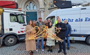 Fünf Personen stehen auf gepflastertem Platz zwischen zwei weißen Fahrzeugen und halten eine beleuchtete Dekoration mit Sternen.
