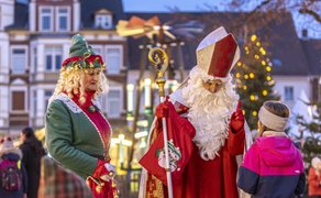 Person in rotem Bischofsgewand mit Mitra und Stab spricht mit Kind in pinker Jacke, daneben steht eine Person in grünem Elfenkostüm mit rotem Schal, im Hintergrund weihnachtlich beleuchtete Fachwerkhäuser.