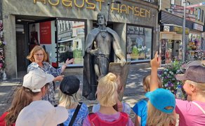 Eine Stadtführerin erklärt einer Gruppe von Kindern eine Bronzestatue in der Peiner Innenstadt; mehrere Kinder hören zu und zeigen interessiert darauf, im Hintergrund sind Geschäfte und Wegweiser zu sehen.