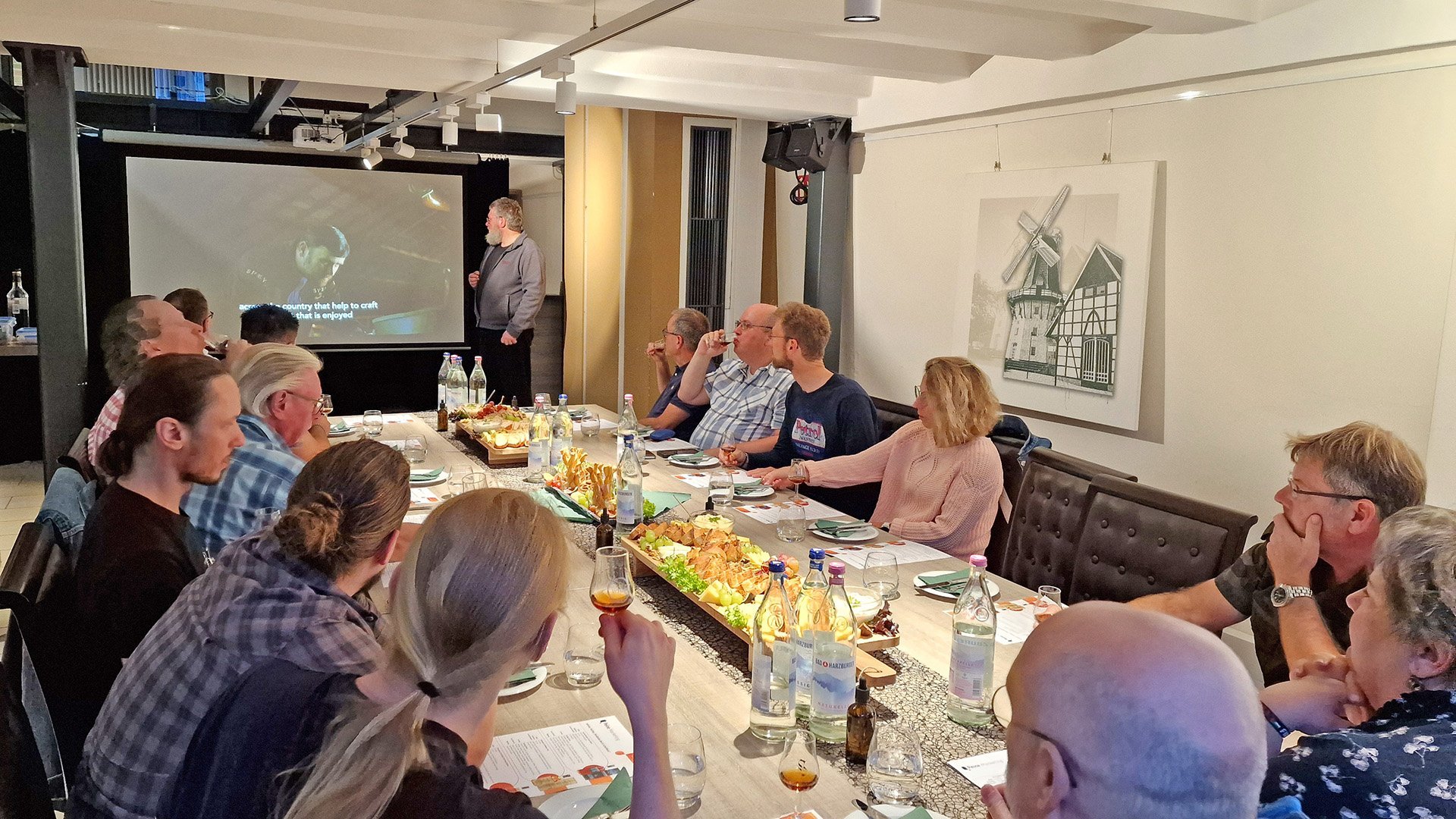 Eine Gruppe von Menschen sitzt gemeinsam an einem Tisch mit Käseplatte und Gläsern und verfolgt eine Präsentation.