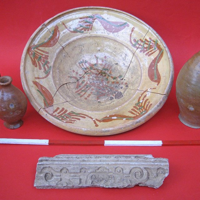 Vier archäologische Keramikgefäße und ein verziertes Steinfragment auf rotem Untergrund.