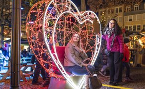 Zwei Frauen vor einem großen, herzförmigen, rot-weiß beleuchteten Deko-Objekt auf einem belebten Weihnachtsmarkt bei Nacht.