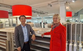 Ein Mann im Anzug und eine Frau im Mantel stehen in einem Baumarkt vor Regalen mit Fliesenmustern.