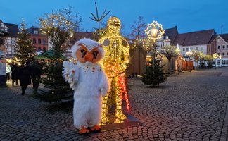 Uhlinchen steht auf dem weihnachtlich beleuchteten Marktplatz in Peine neben einer leuchtenden Ritterfigur, umgeben von Tannenbäumen und Weihnachtsbeleuchtung.