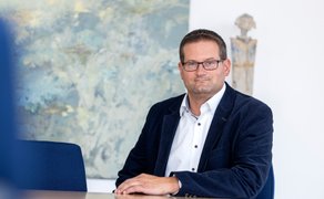 Ein Mann in einem weißem Hemd und dunkelblauem Sakko sitzt an einem Tisch.