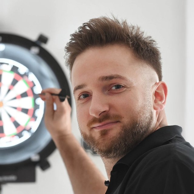 Mann in schwarzem Poloshirt hält Dartpfeil vor beleuchteter Dartscheibe und schaut in die Kamera.