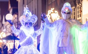 Zwei Personen in weißen, leuchtenden Kostümen mit Kronen und Umhängen halten sich an den Händen, umgeben von weihnachtlicher Straßenbeleuchtung.