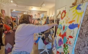 Frau malt mit Pinsel auf große Leinwand mit bunten Blumen, Sonne und Herzen in einem Raum mit Kleidung und weiteren Personen.